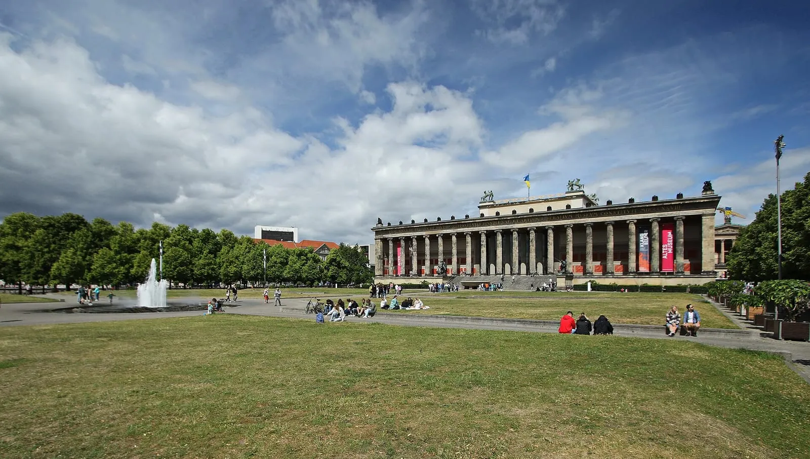 La colonnade ionique de l'Altes Museum vue depuis la pelouse du Lustgarten, avec la fontaine à gauche et des visiteurs assis sur l'herbe