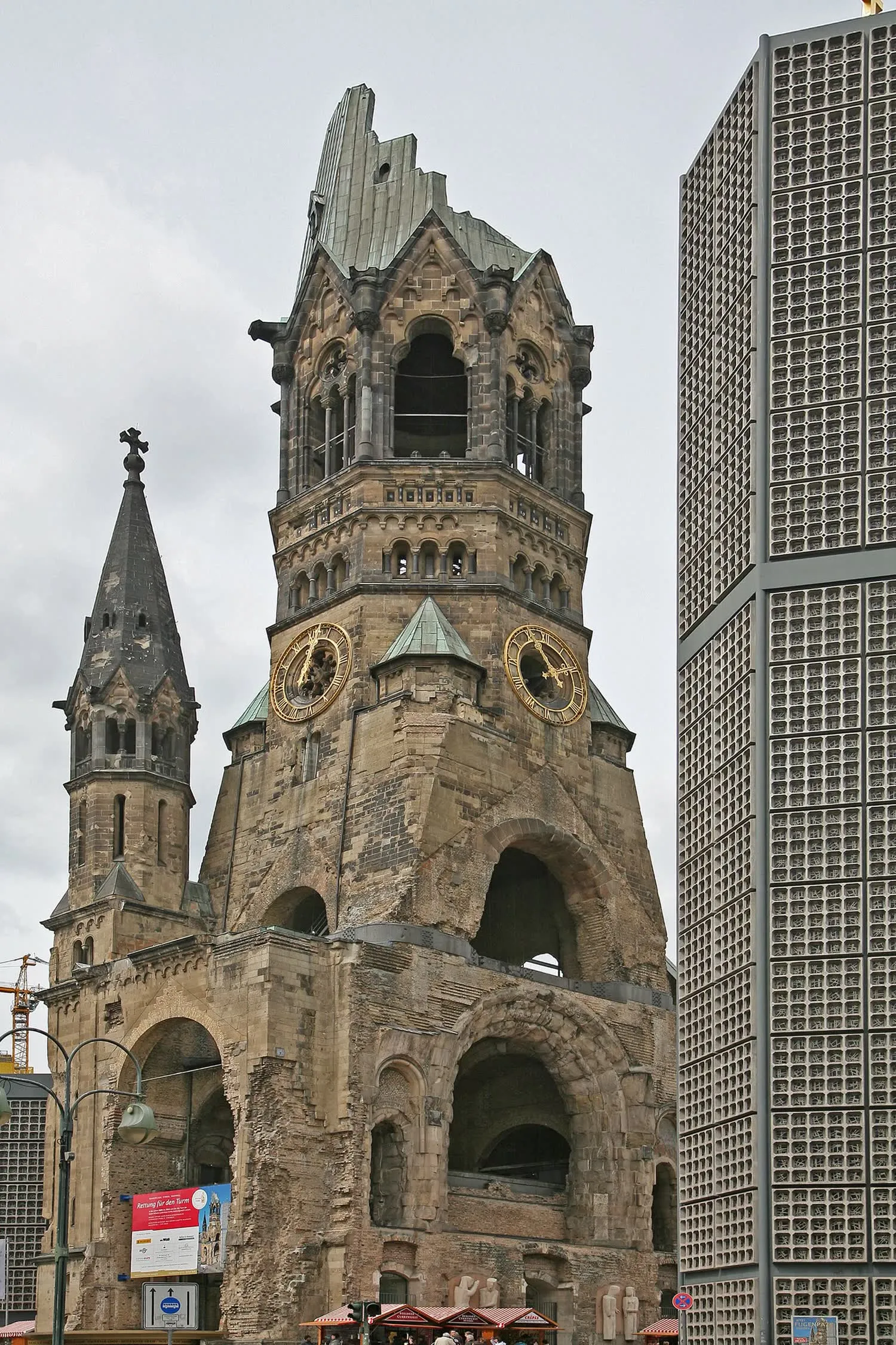 La tour ruinée de l'église du Souvenir, sommet arraché et cadrans d'horloge intacts, avec la façade en alvéoles de béton de la nouvelle église sur la droite