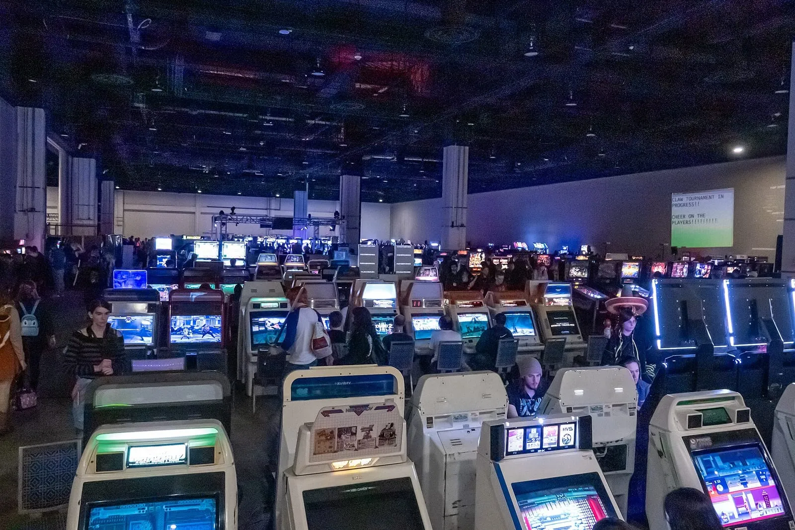Rangées de bornes d'arcade éclairées par des néons, avec des joueurs assis devant les écrans dans une grande salle de jeux