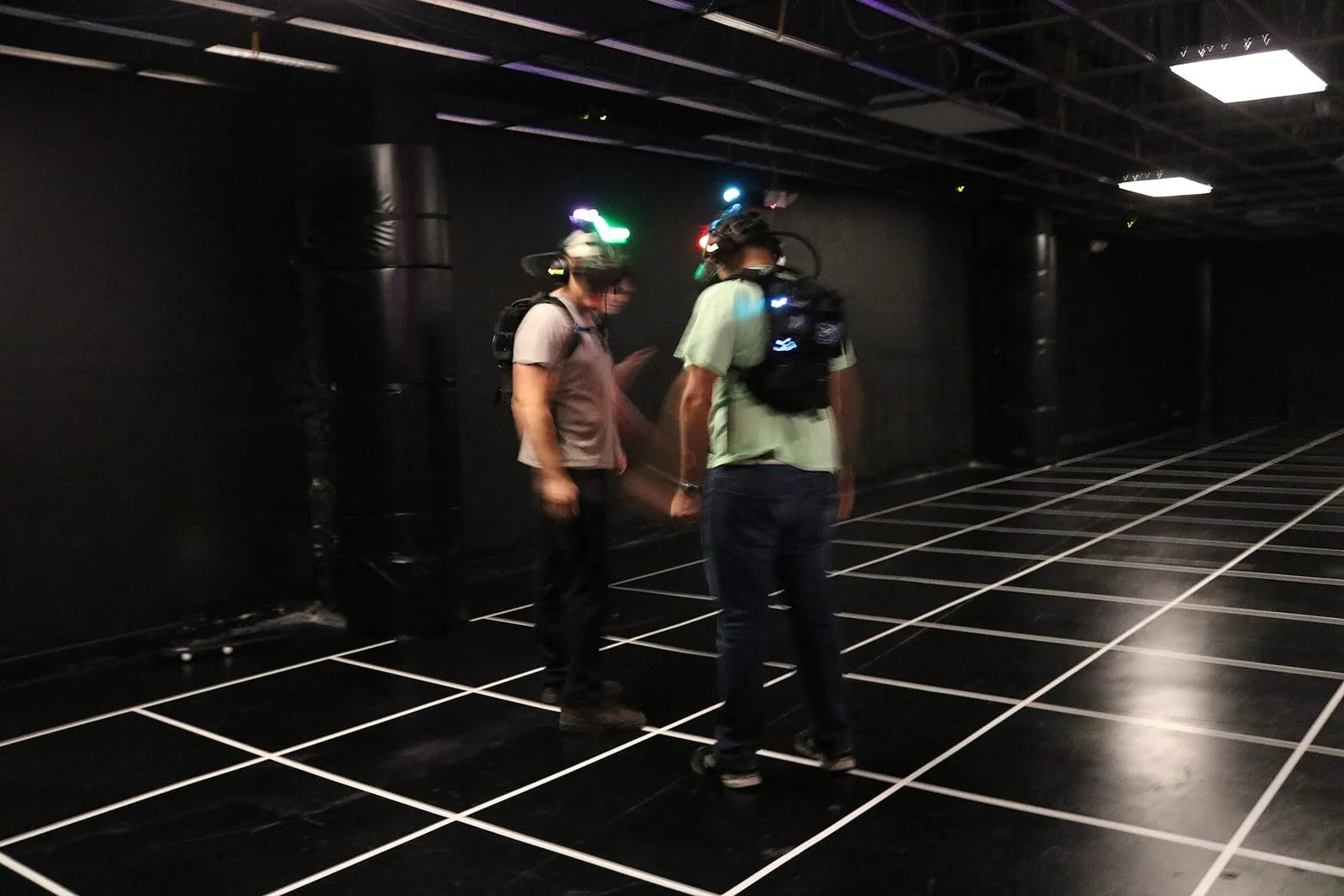 Deux joueurs équipés de casques de réalité virtuelle et de sacs à dos de suivi, debout sur le sol quadrillé d'une arène de free-roam