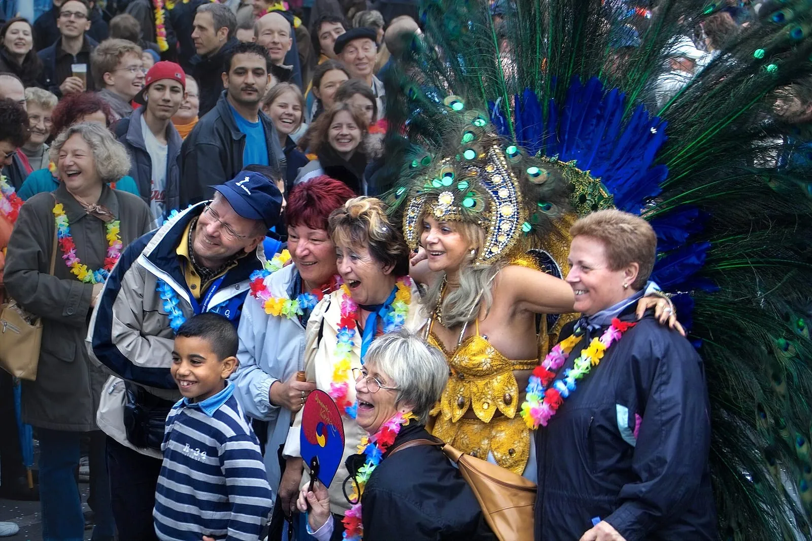 Participante au Carnaval des cultures de Berlin : costume doré à paillettes et haute coiffe de plumes de paon, entourée de spectateurs au bord du défilé