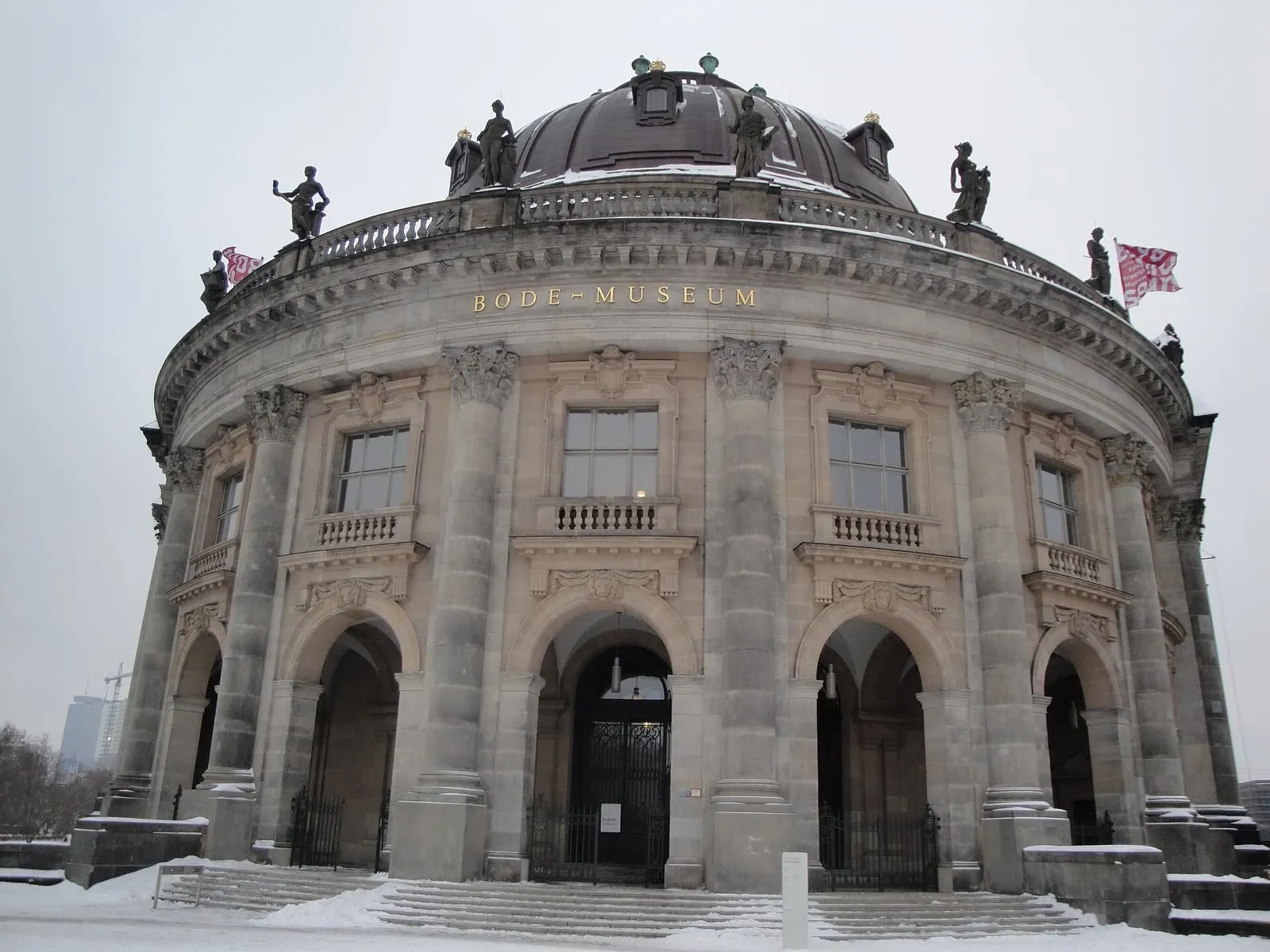 La façade arrondie du Bode Museum sous la neige, escaliers et coupole blanchis, sous un ciel d'hiver uniformément gris