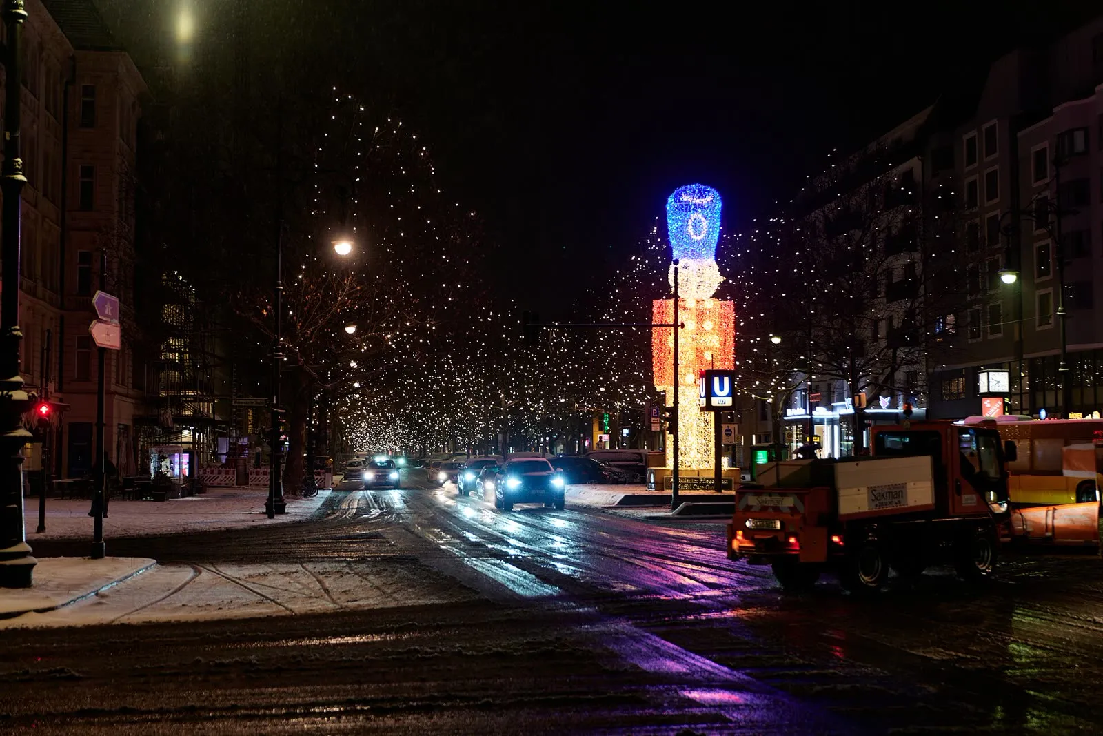 Le Kurfürstendamm par une nuit d'hiver après une chute de neige, chaussée luisante, voitures et illuminations accrochées aux arbres, camion de déneigement au premier plan