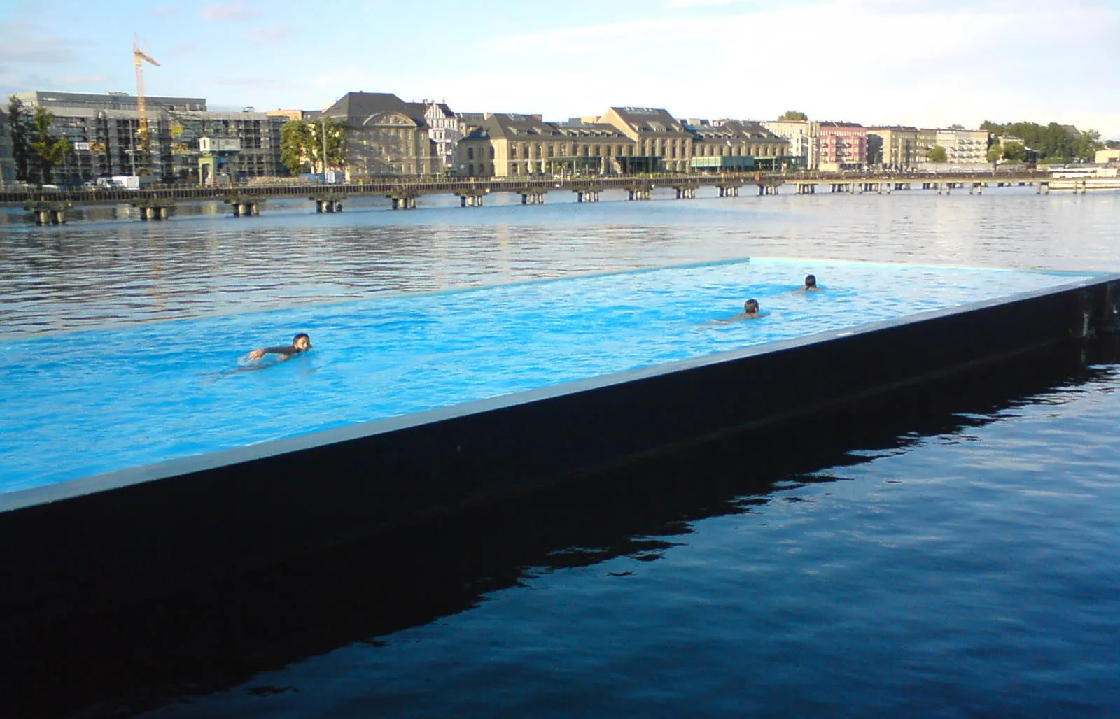 Le Badeschiff, piscine rectangulaire à bord noir amarrée sur la Spree à Berlin, trois nageurs dans l'eau, immeubles de la rive en arrière-plan