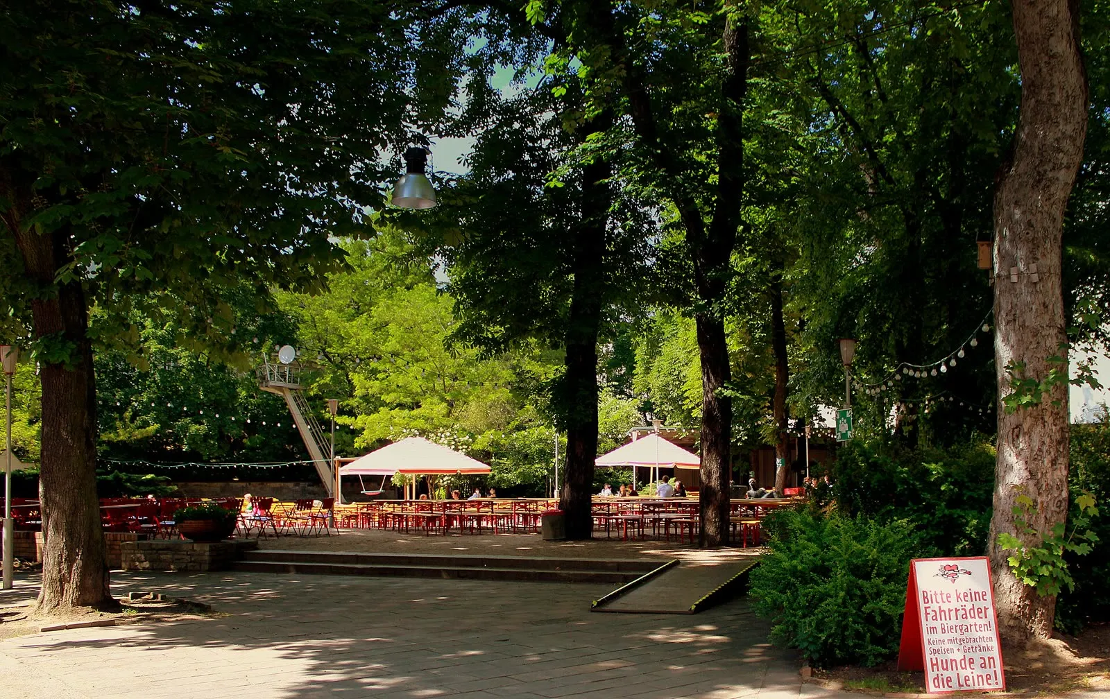Tables et bancs en bois d'un Biergarten berlinois sous de grands arbres, parasols et guirlandes lumineuses, panneau rouge interdisant les vélos dans le jardin