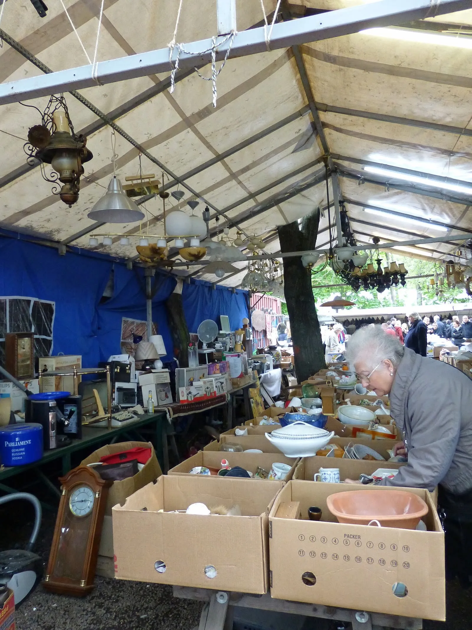 Stand de brocante sous bâche au marché aux puces de Mauerpark, luminaires et lustres anciens suspendus, vaisselle et pendule posées dans des caisses en bois