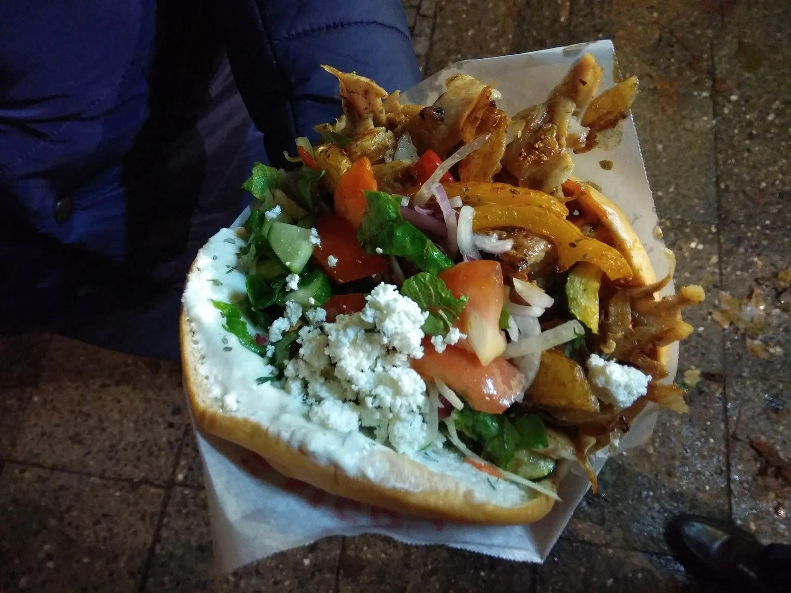 Un döner kebab garni de salade, de sauce blanche, de fromage et de frites, tenu dans son emballage papier