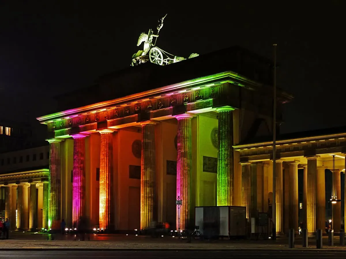 La porte de Brandebourg illuminée de nuit par des projections multicolores du Festival of Lights, quadrige éclairée en vert au sommet et promeneurs devant les colonnes