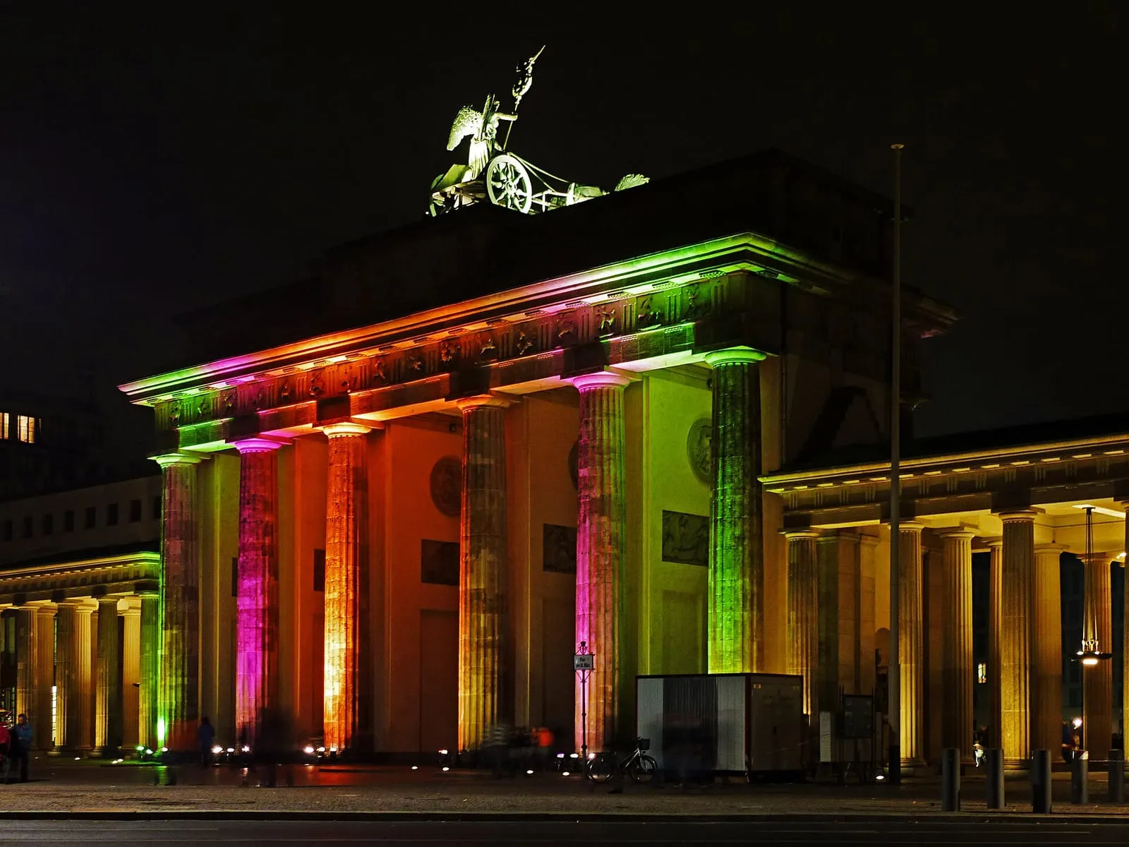 La porte de Brandebourg illuminée de nuit par des projections multicolores du Festival of Lights, quadrige éclairée en vert au sommet et promeneurs devant les colonnes