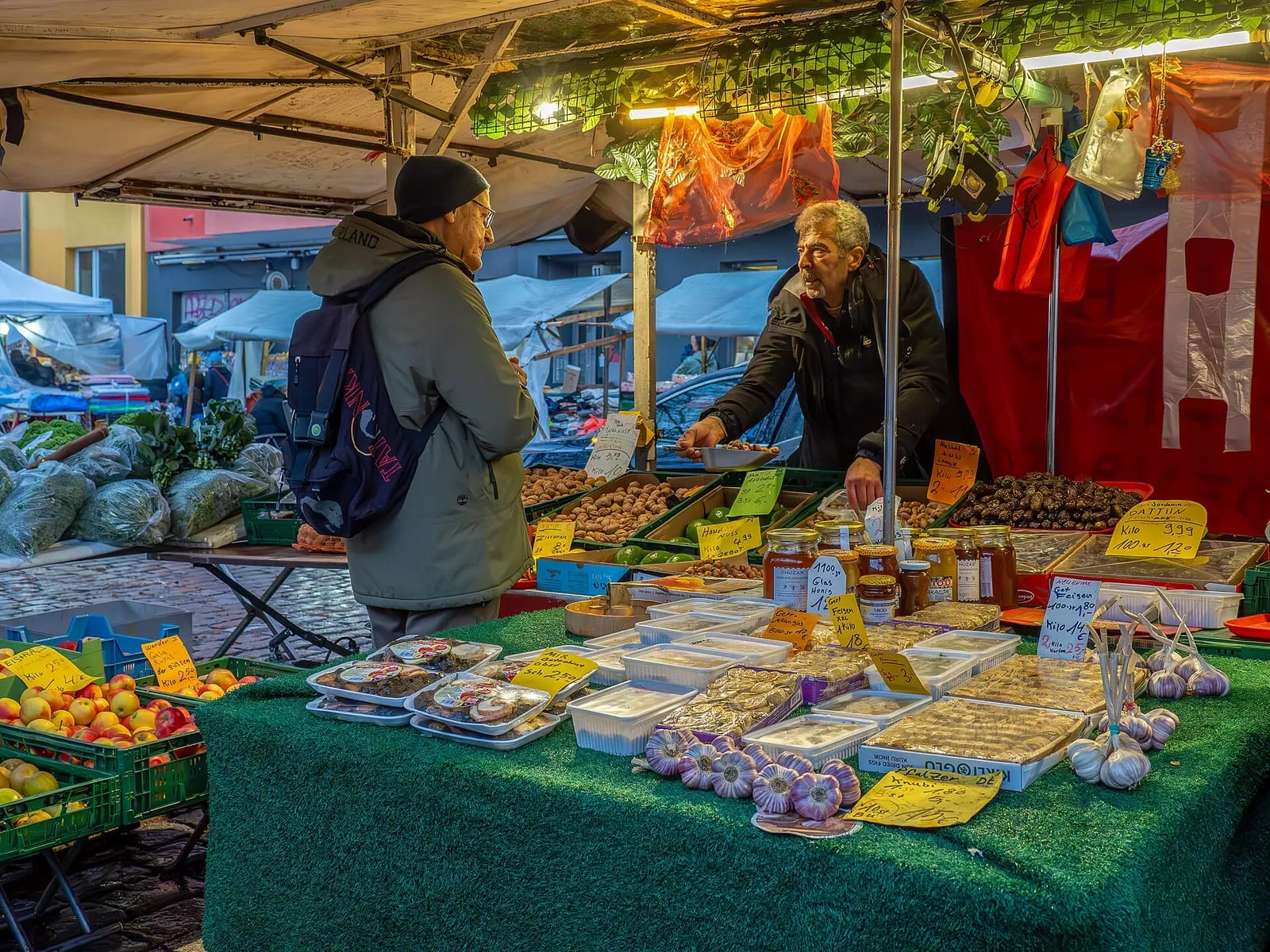 Étal d'un marché en plein air à Berlin, avec pommes vertes en cageots, tomates, champignons, fromages et conserves, vendeur et client devant la marchandise