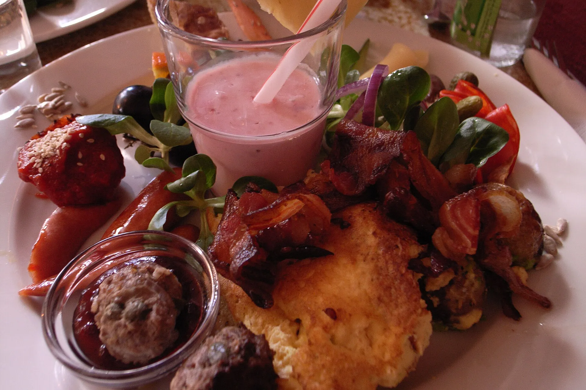 Assiette de brunch garnie de pommes de terre rôties, de bacon, de salades, d'olives et de condiments, avec un verre de smoothie rose