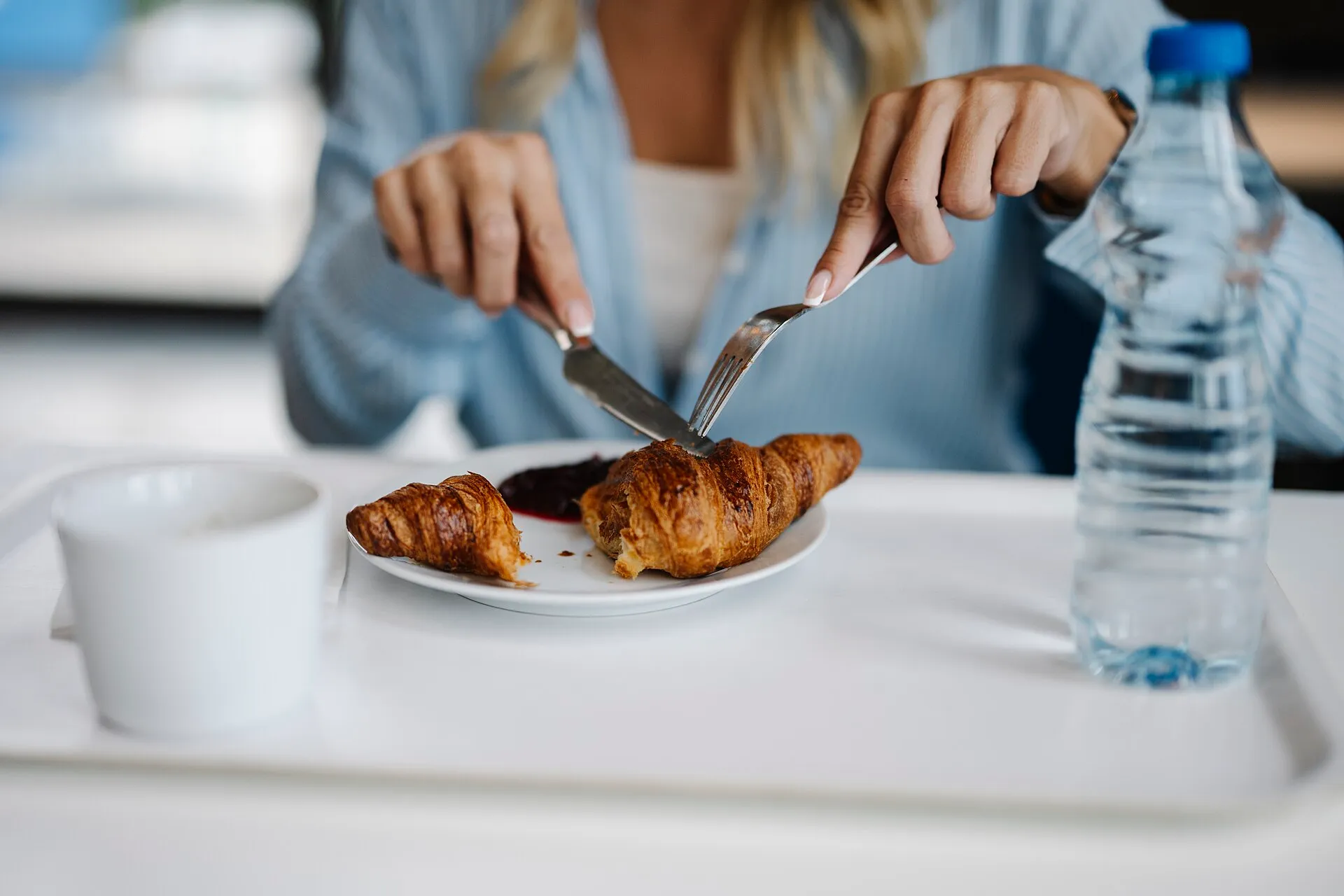 Une personne coupe un croissant posé sur une assiette avec de la confiture, à côté d'une tasse de café