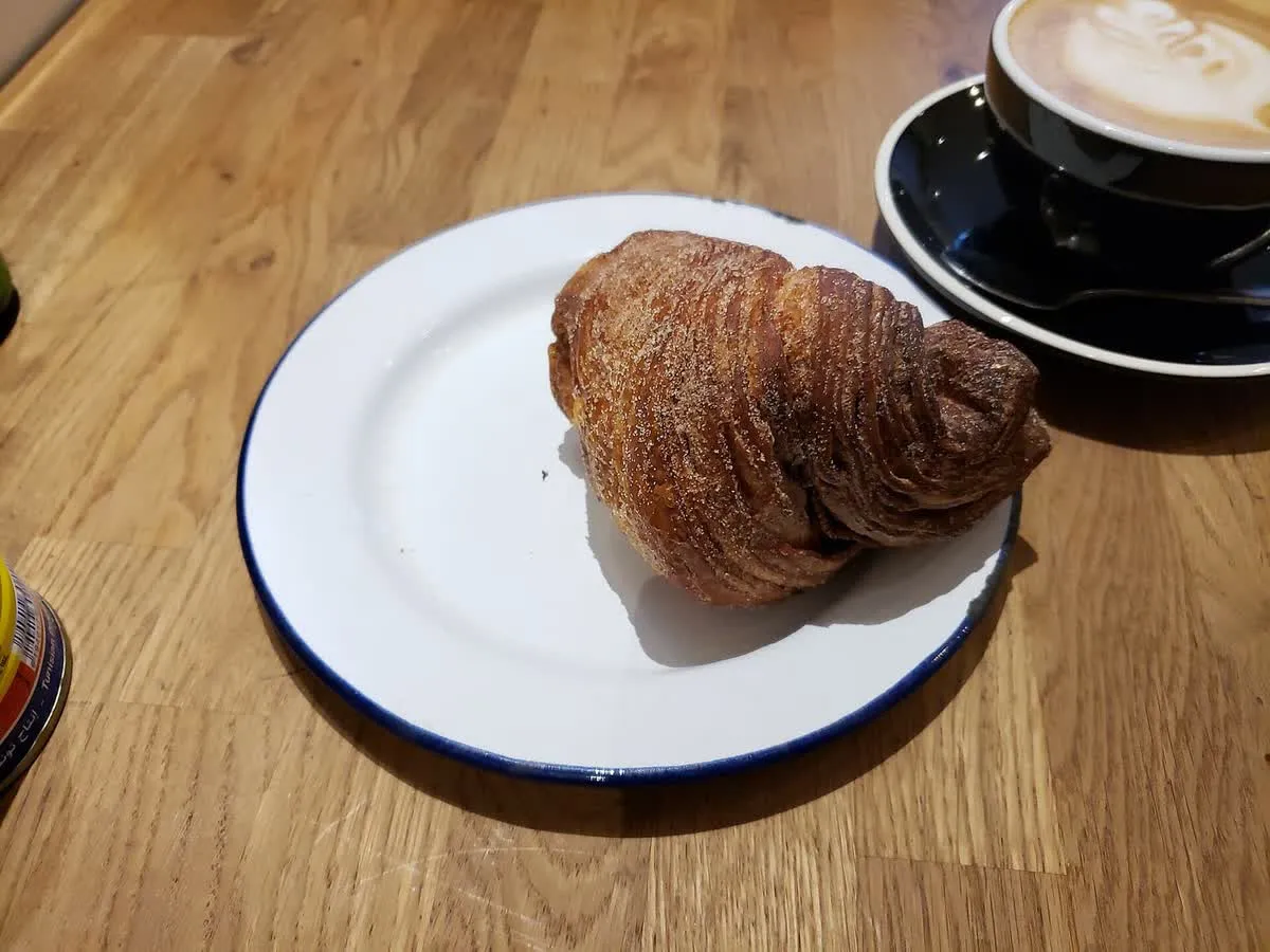 Grosse viennoiserie roulée saupoudrée de sucre sur une assiette blanche, accompagnée d'un café latte