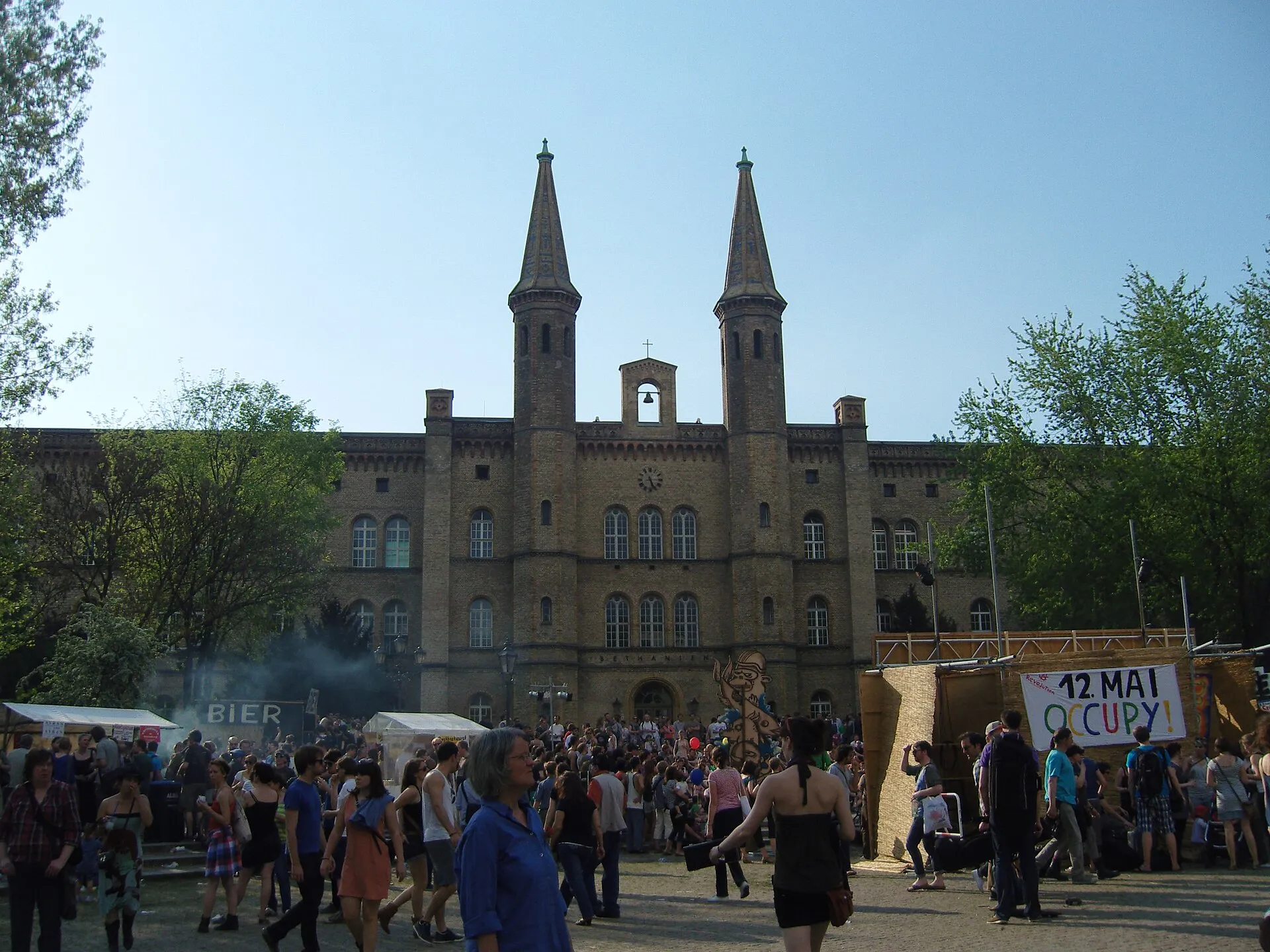 Foule rassemblée sur le parvis du Bethanien, à Kreuzberg, lors d'une fête de mai : stands, bannières et promeneurs sous un ciel dégagé