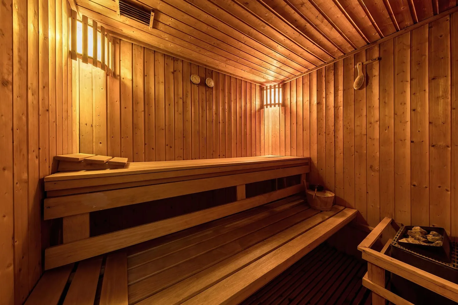 Intérieur d'un sauna moderne en bois clair, avec deux niveaux de bancs et un poêle garni de pierres