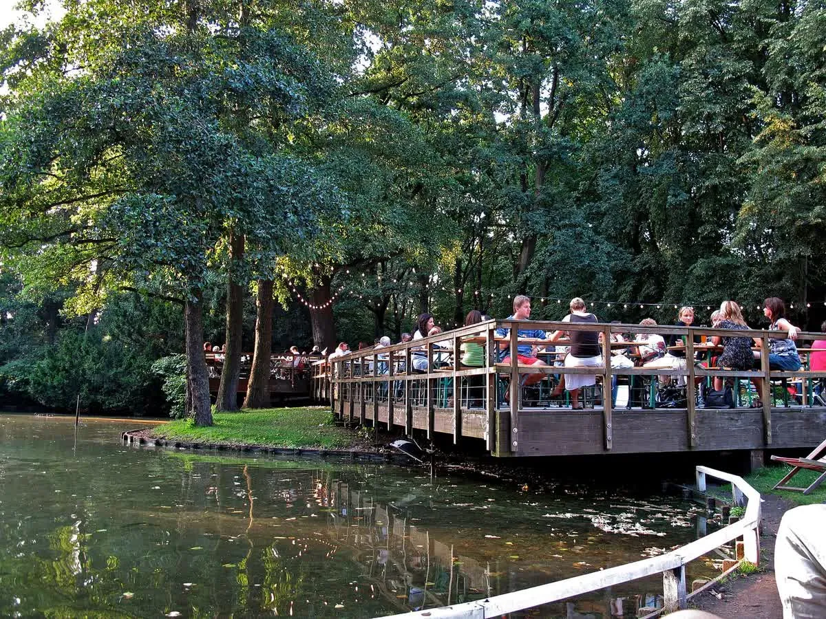 La terrasse en bois du Café am Neuen See, posée sur l'eau du Neuer See, avec ses tables et ses clients installés sous les arbres