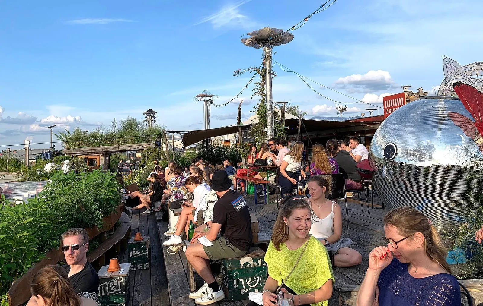 La terrasse en bois du Klunkerkranich, à Neukölln, remplie de jeunes gens assis sous les plantes, devant une grosse boule à facettes, avec les toits de Berlin en arrière-plan