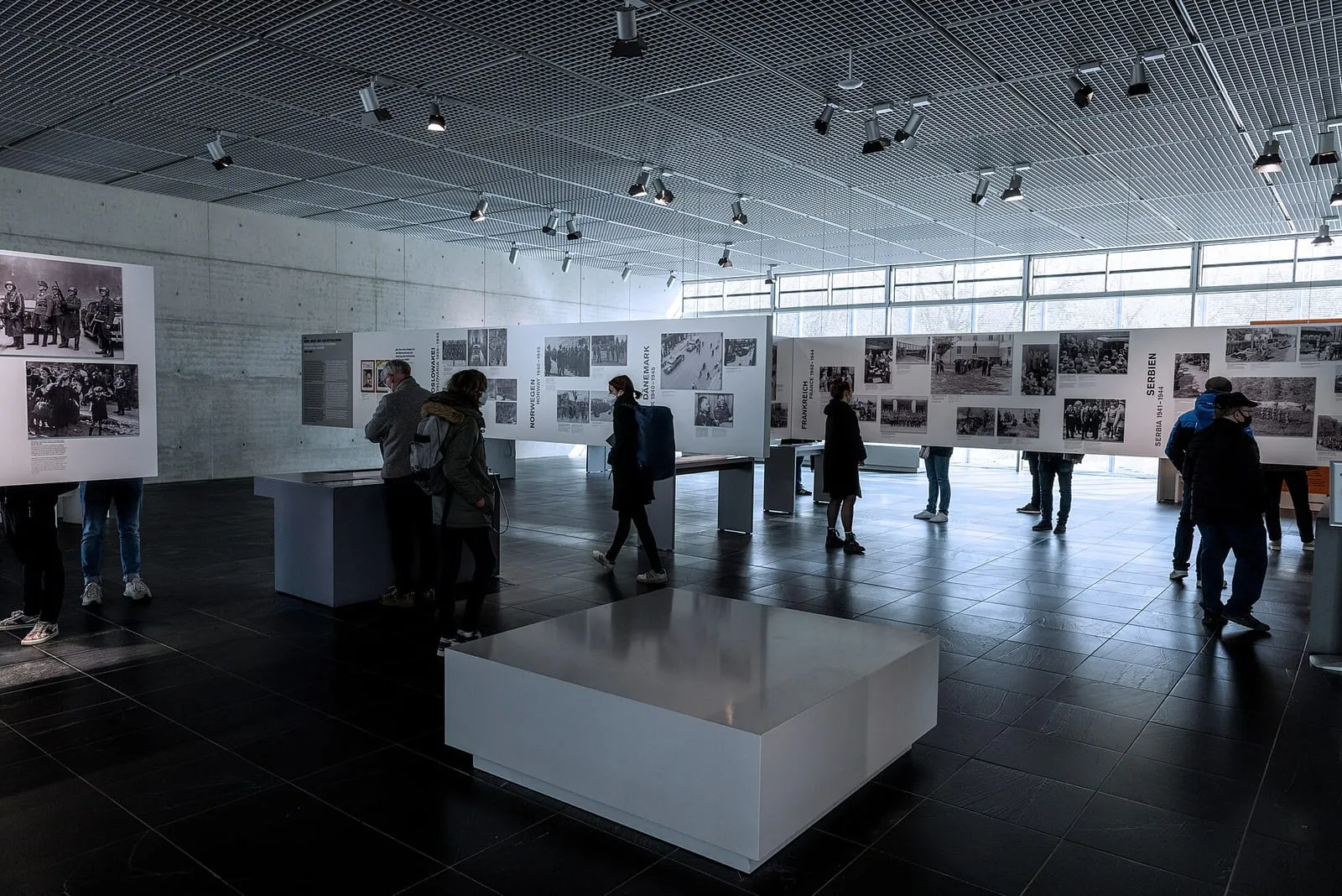 La salle de l'exposition permanente : panneaux de photographies historiques, pupitres de documents et plafond à grille avec projecteurs
