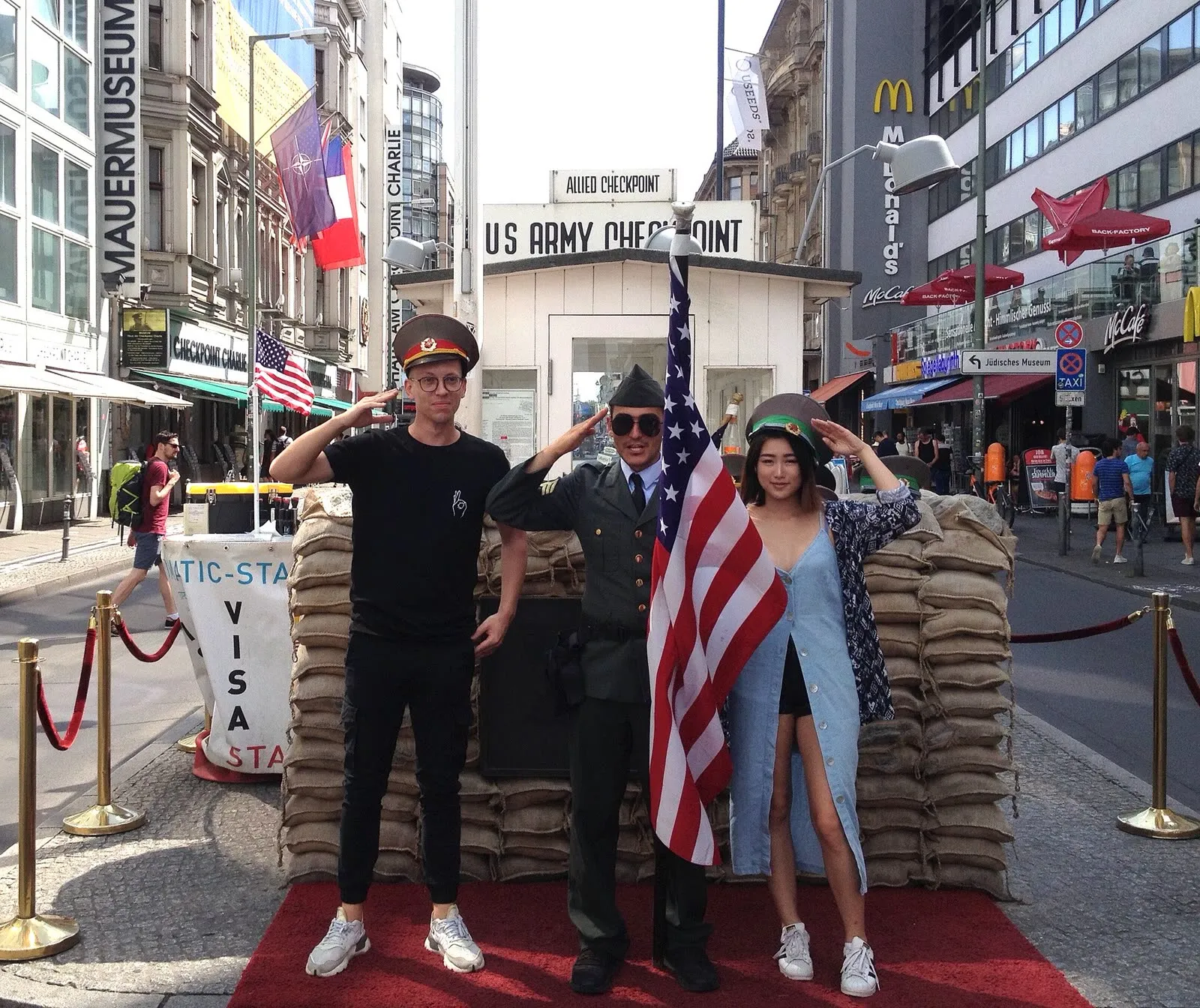 Deux touristes posent devant la cabane du Checkpoint Charlie, entre les sacs de sable, le drapeau américain et un figurant en uniforme
