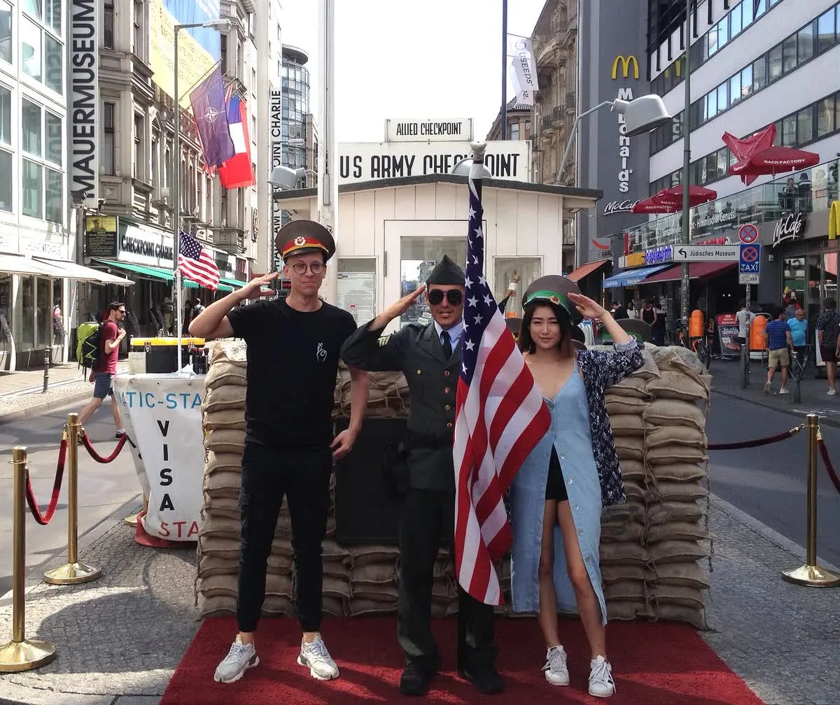 Deux touristes posent devant la cabane du Checkpoint Charlie, entre les sacs de sable, le drapeau américain et un figurant en uniforme