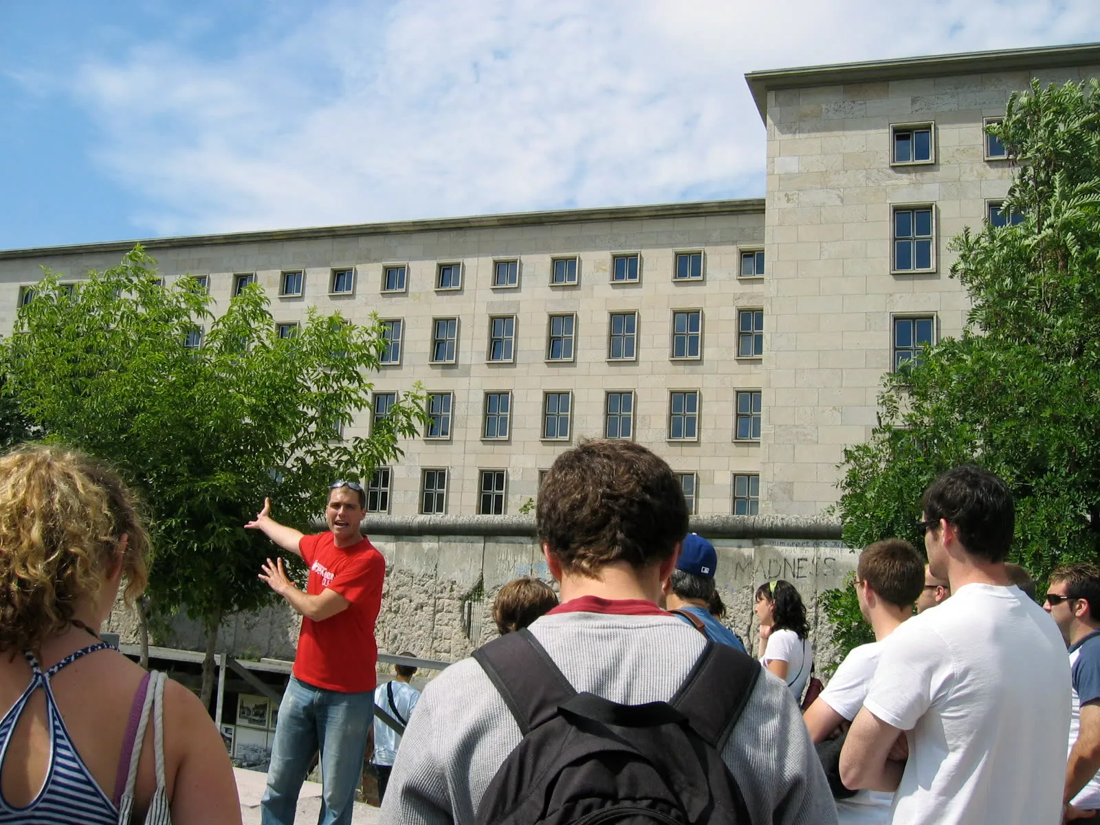 Un guide en chemise rouge, bras tendu, commente une façade devant un groupe de visiteurs, un vestige du mur de Berlin au premier plan